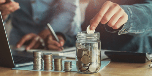 A hand places a coin into a jar that is half full while other coins stand stacked next to the jar